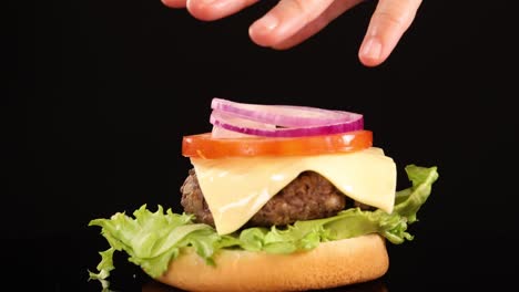 hands layer tomato, onion, cheese, and lettuce onto a beef patty and bun under bright studio lighting, finishing with a sesame seed top bun