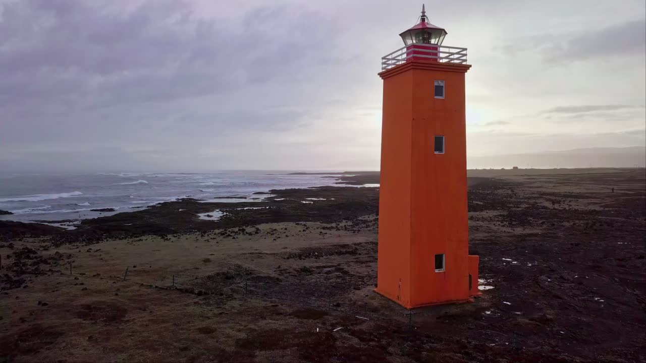 Faro arancione retroilluminato da un sole al tramonto in una giornata nuvolosa sulla costa islandese
