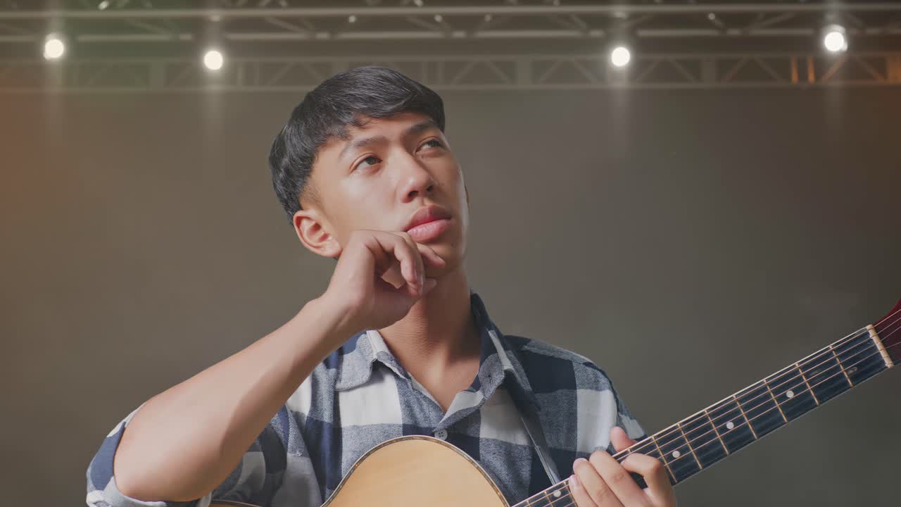 기타를 들고 있는 아시아 십대가 무언가에 대해 생각하고 주위를 둘러보며 무대에서 지표손가락을 들어 올린다.