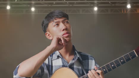 primer plano de un adolescente asiático con una guitarra pensando en algo y mirando a su alrededor luego levantando su dedo índice en el escenario