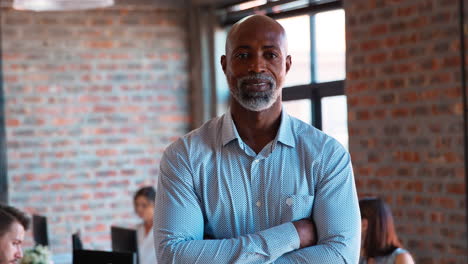 Portrait-Of-Smiling-Mature-Businessman-Standing-In-Busy-Office-With-Colleagues-Working-In-Background