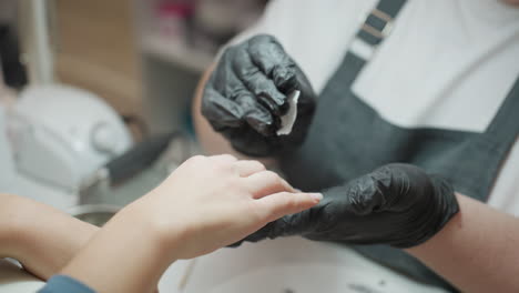 nail technician in black glove gently cleaning client hand with wipe during manicure session, showing care and precision in professional nail salon environment with visible equipment in background
