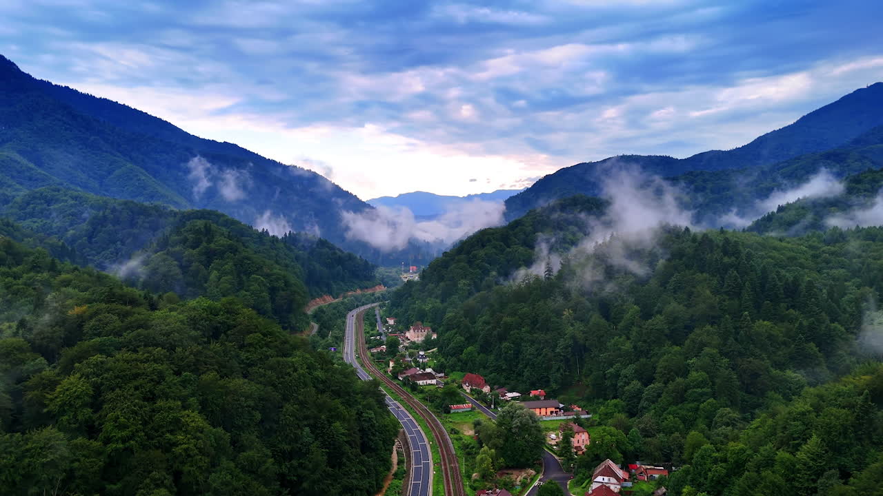 Village in the valley between the green mountains. Drone footage above the highway and railway crossing the landscape. Romania