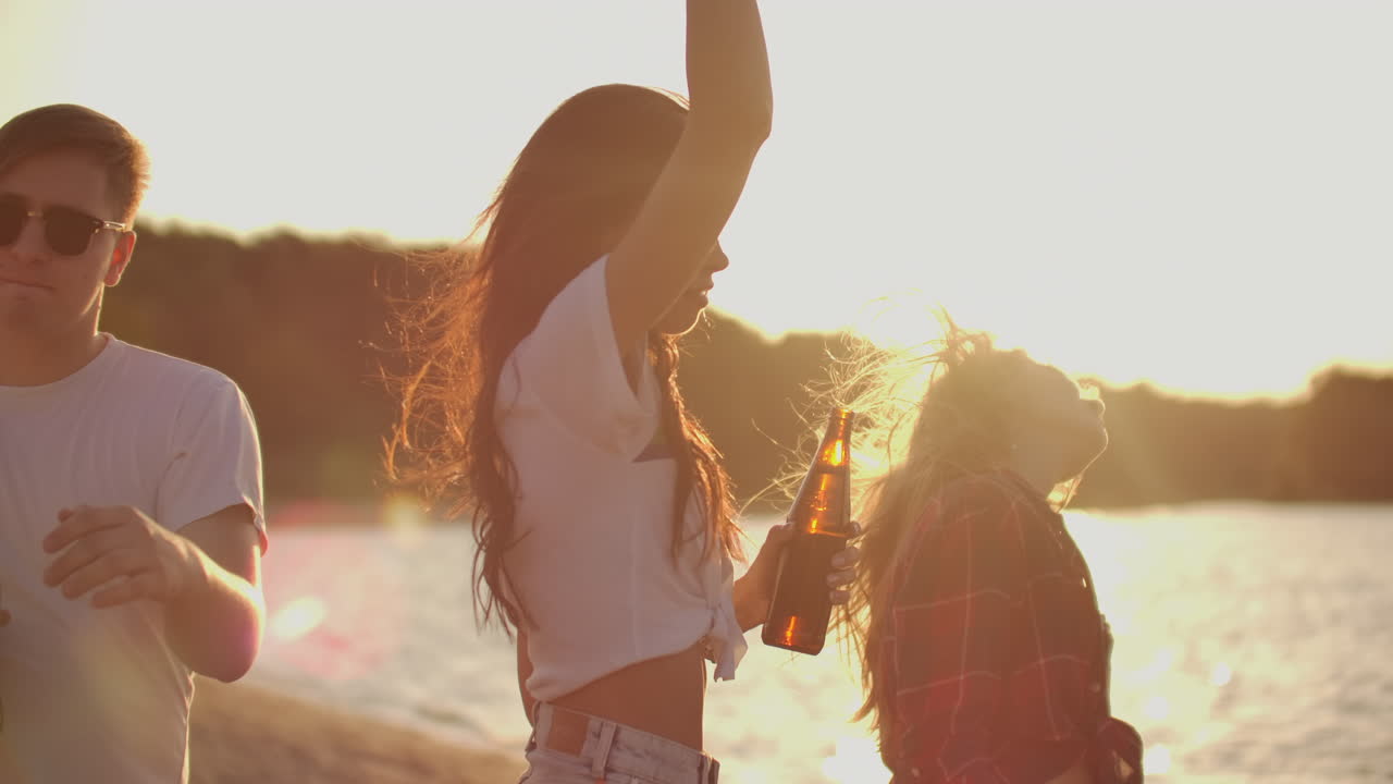 두 소녀와 소년은 야외 파티에서 맥주와 좋은 분위기로 생일을 축하합니다. 그들은 강변 근처의 여름 저녁에 춤을 니다.