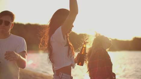 two girls and boy celebrate a birthday on the open air party with beer and good mood. they dance in the summer evening near the river coast.