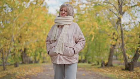 elderly woman in light pink puffer jacket and thick cream scarf walking on leaf covered path in colorful autumn park, hands in pockets, looking thoughtful under golden yellow tree canopy