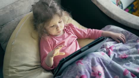 Porträt-Eines-Hübschen-Kleinen-Mädchens,-Eines-Kindes,-Das-Auf-Dem-Sofa-Unter-Der-Decke-Liegt-Und-Auf-Einem-Tablet-Computer-Spielt