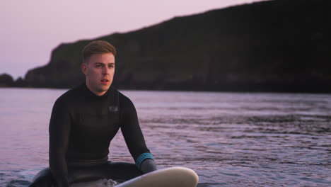 man wearing wetsuit sitting and floating on surfboard on calm sea