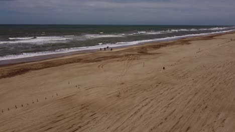 Aerial-closing-up-view-with-a-wide-angle-beach-with-people-riding-horses-on-the-beach-sand-on-a-mild-evening-at-Mar-de-las-Pampas,-Argentina