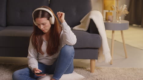 Mujer-Con-Auriculares-Inalámbricos-Sentada-En-El-Suelo-En-Casa-Transmitiendo-Música-Desde-Un-Teléfono-Móvil-12