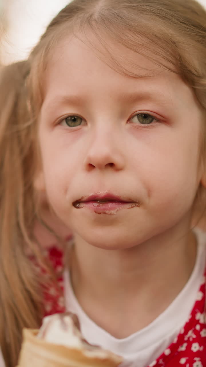 Preschooler girl with ponytails eats ice cream with pleasure sitting on wooden bench closeup. Kid bites cold dessert at sunset. Delicious sundae snack