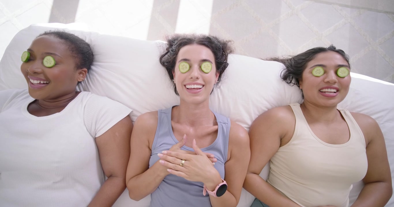 Drie vriendinnen liggen op bed, de middelste vrouw legt komkommerschijfjes voor een thuis-spa-sessie.