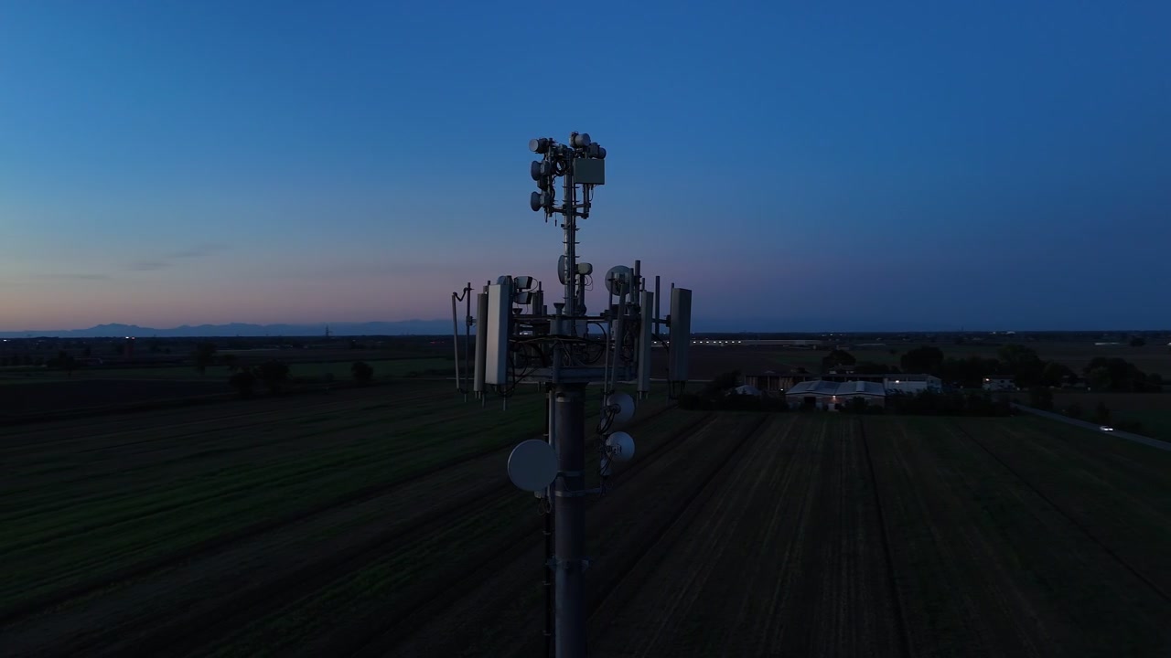 Torre di telecomunicazioni con antenne a pannello settoriali, parabole a microonde, unità radio e apparecchiature di supporto sopra la Pianura Padana nel nord Italia durante un'orbita ravvicinata del drone con effetto parallasse.