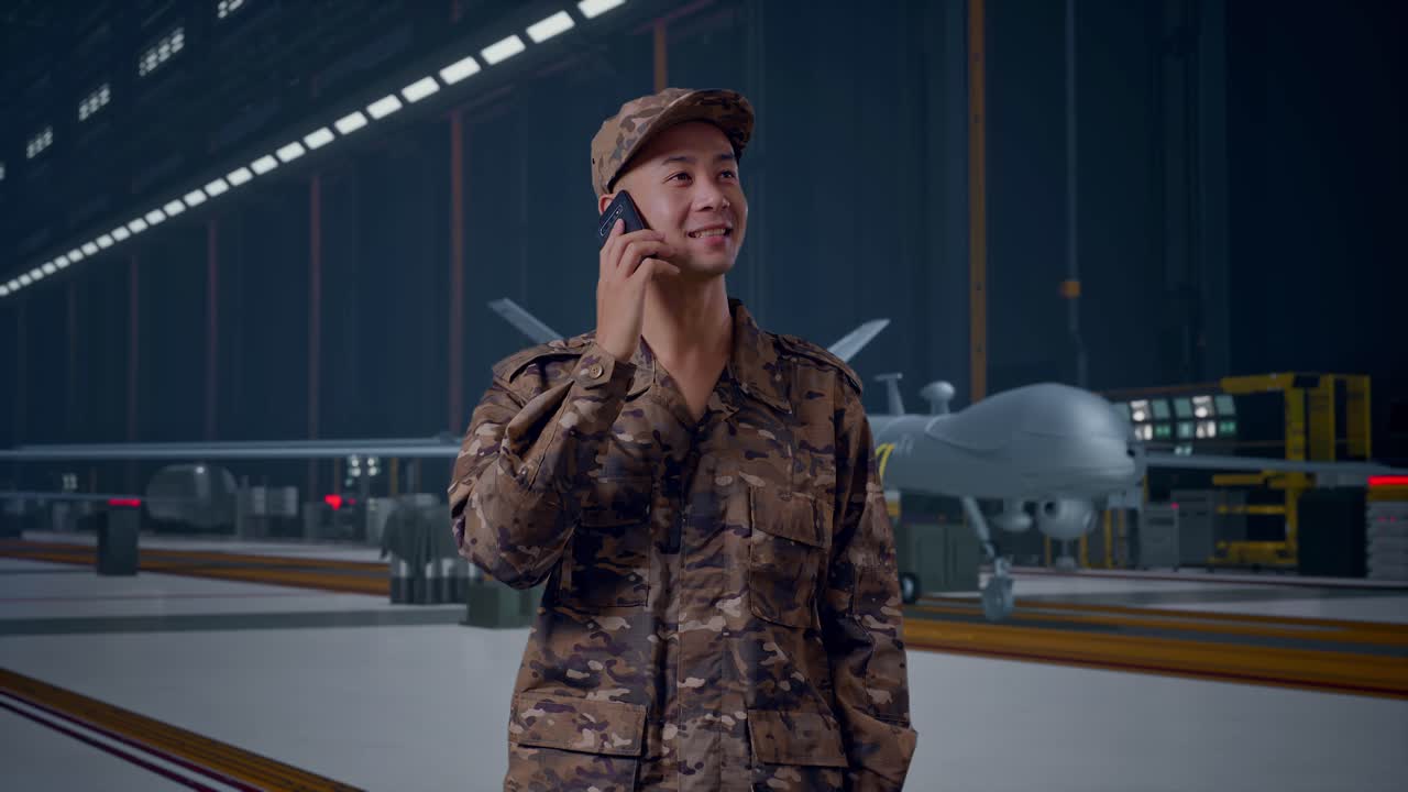Asian Man Soldier Talking On Smartphone And Looking Around While Standing With Modern Military Drone In Maintenance Hangar