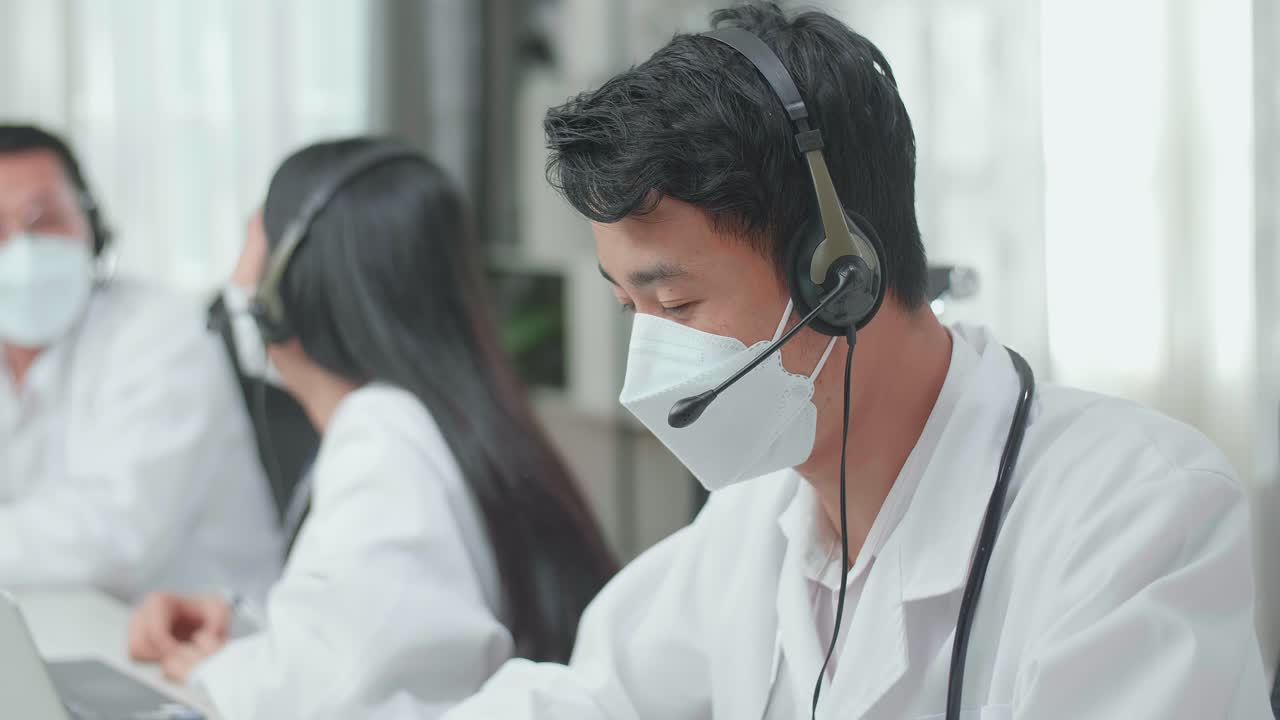 Close Up Of An Asian Man Doctor Wearing Headsets And Masks Working As Call Center Agent Speaking To Customers On The Call And Typing While His Colleagues Are Talking During Working At The Office