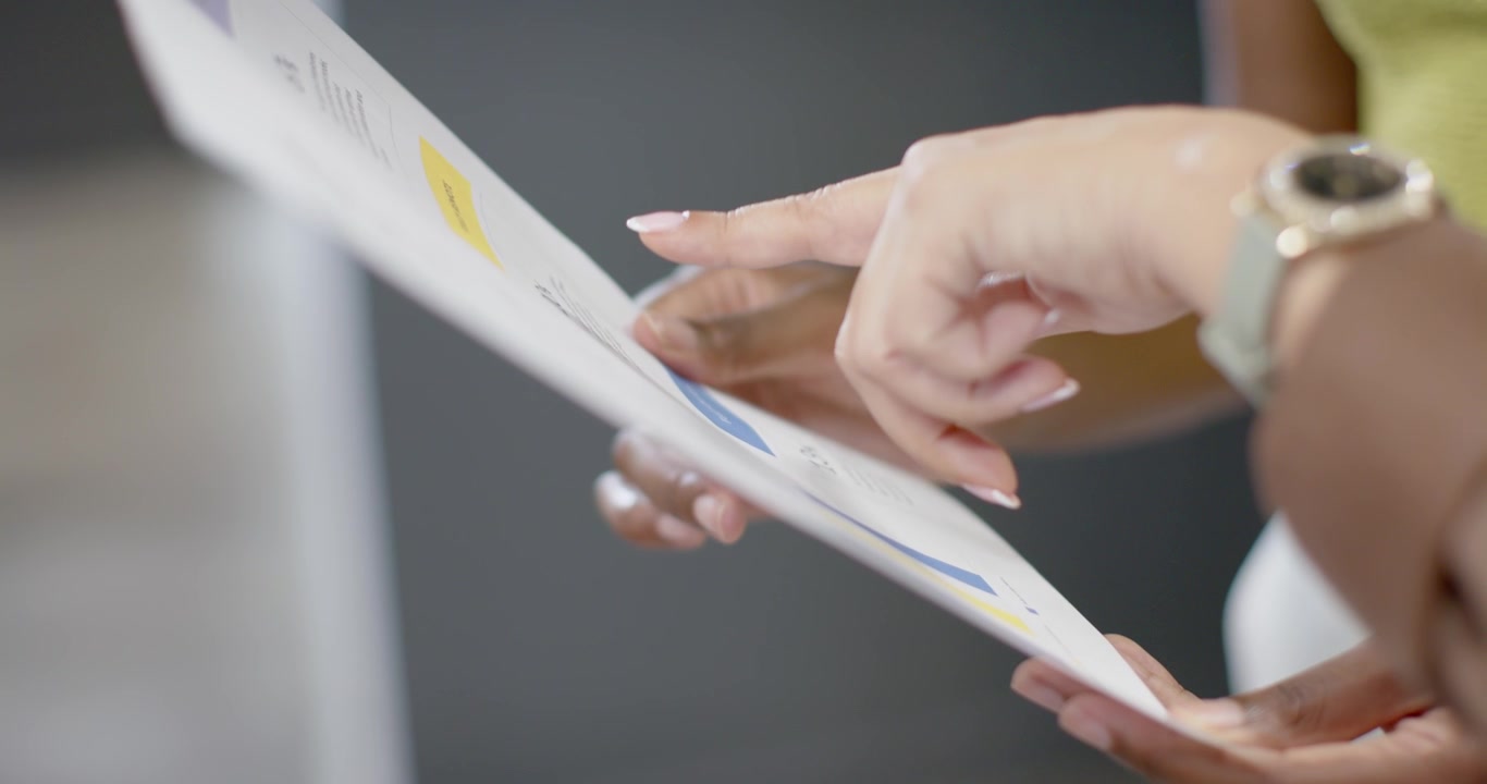 Diverse female team checking report at desk, holding edge while peer bringing hand pointing charts