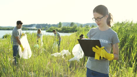 Young-Pretty-Woman-Standing-With-Alist-Of-Volunteers-In-Front-Of-The-Camera-And-Smiling-While-Students-Volunteering-Behind-And-Cleaning-Up-A-Lake-Area