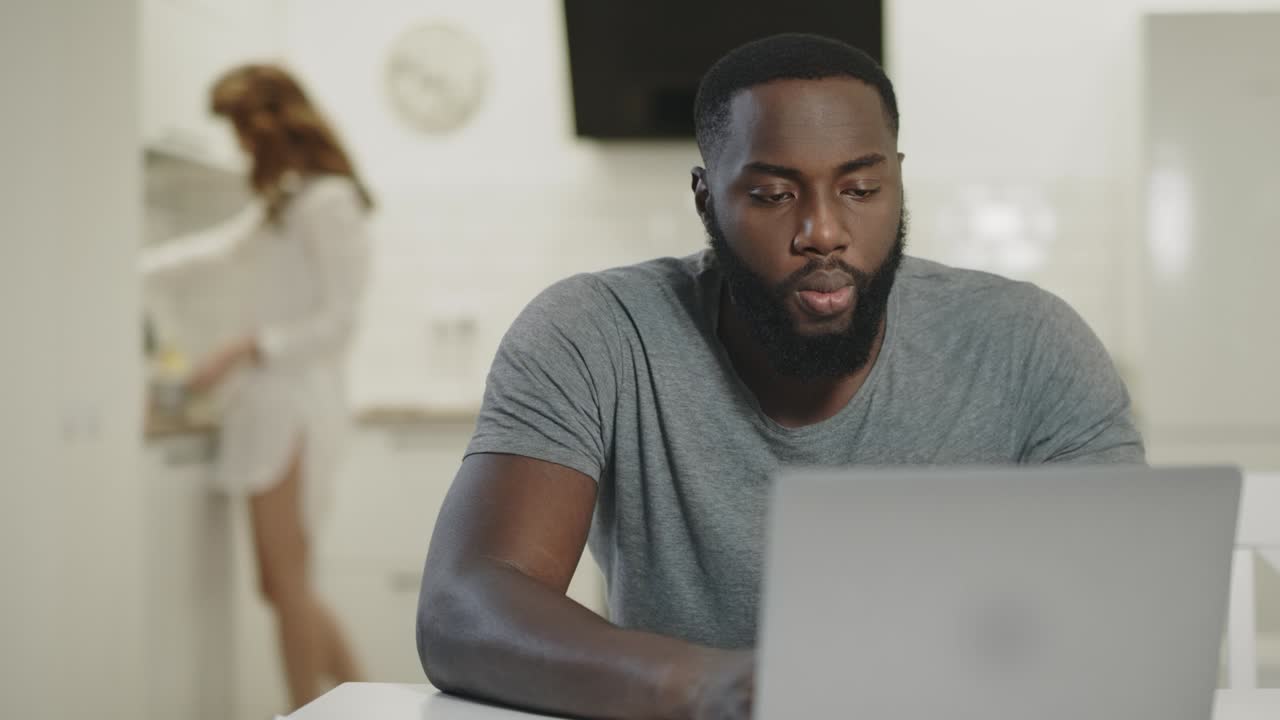 Serious black man working at laptop at open kitchen. Smiling couple drinking tea