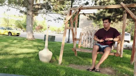 young hispanic man walks to sit on hanging bench in public outdoors park, sunny day