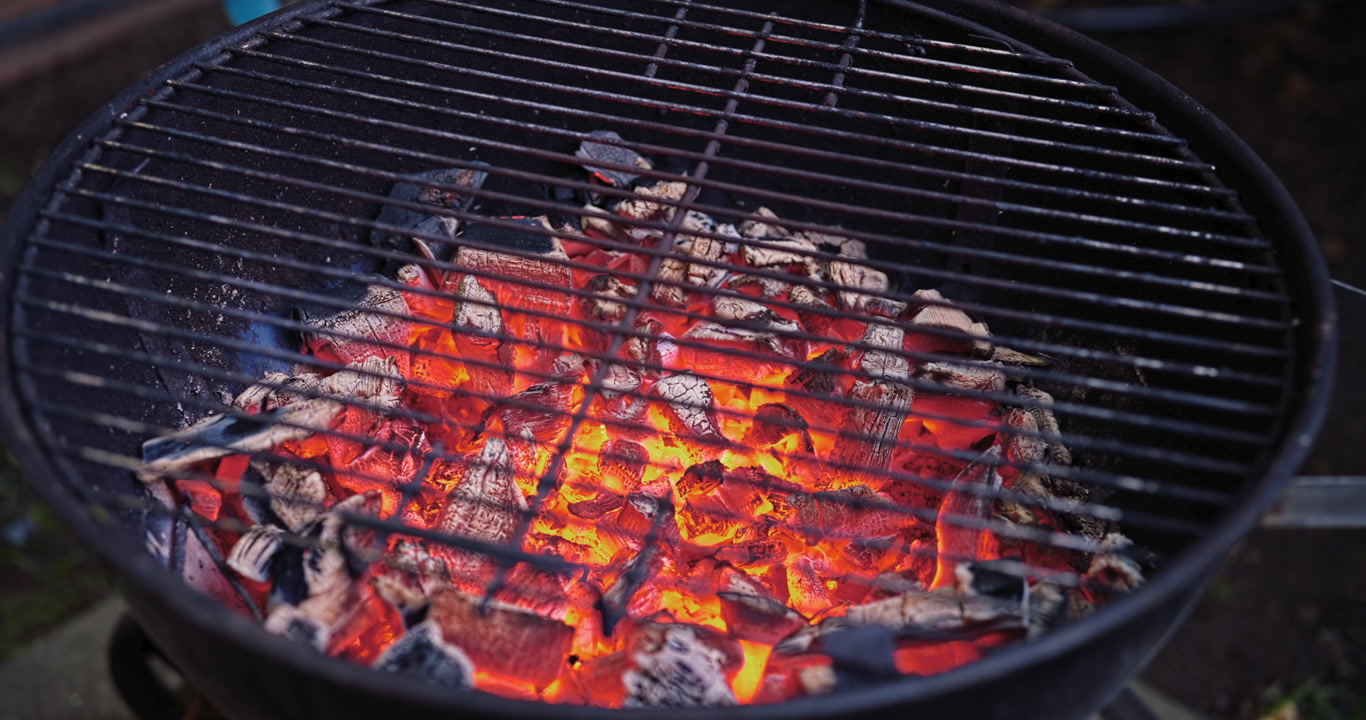 A smooth lateral camera move passes over glowing charcoal embers in a worn backyard barbecue, capturing real heat and the quiet anticipation before cooking begins.