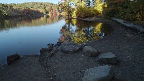 una inclinación lenta comienza desde una pequeña playa con grandes piedras y revela un lago tranquilo con árboles de principios de otoño en la distancia