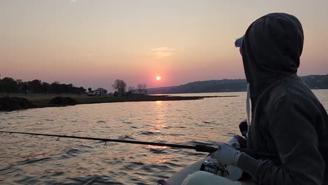 a woman fishing from a small boat on a lake with the sun setting in the distance