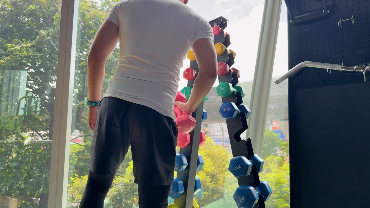 Athletic man selects dumbbell in modern, sunlit gym, preparing for workout near large windows