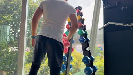 athletic man selects dumbbell in modern, sunlit gym, preparing for workout near large windows