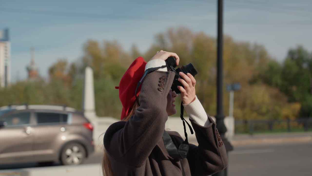 Hvid kvindelig fotograf i rød baret fanger byens skyline med et spejlløst kamera, oprigtige smil og profilbilleder, langt blondt hår, bro og træ som baggrund, solrig blå himmel, stilfuld frakke, rejse.