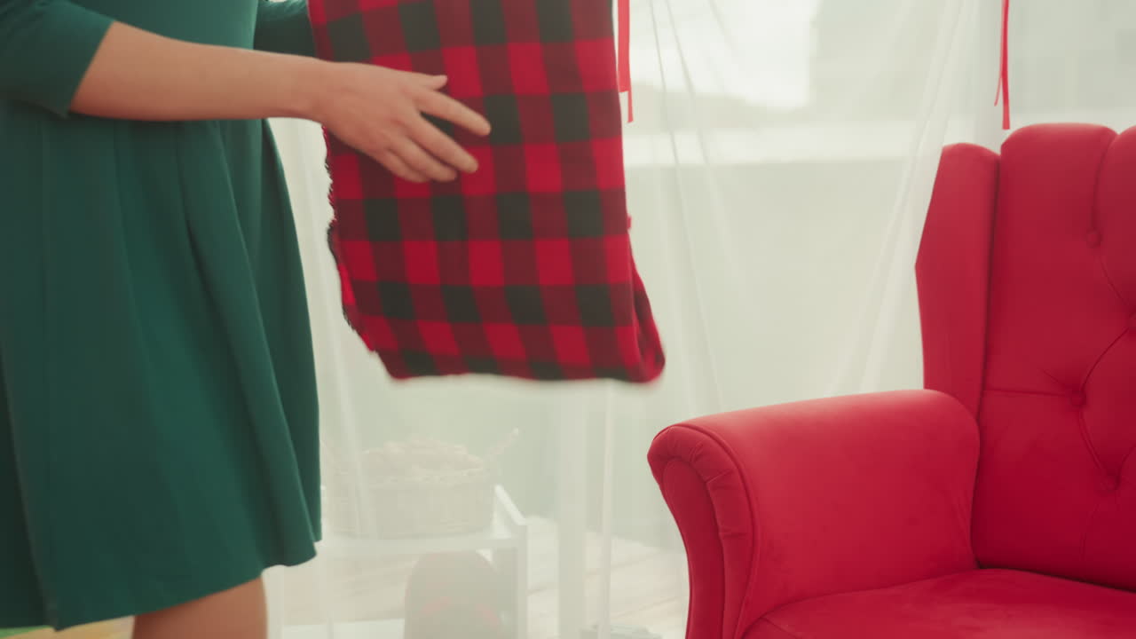 coppia caucasica che regala una coperta a quadri, uomo seduto su un divano rosso trapuntato, donna in abito verde che avvolge una calda coperta a quadri sulle sue spalle, luce naturale brillante attraverso tende leggere, vacanza accogliente