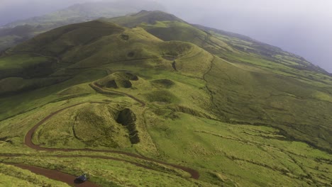 drone dolly view of vulcanic mountains covered in lush green vegetation with dirt road, cloudy weather in são jorge island, the azores, portugal