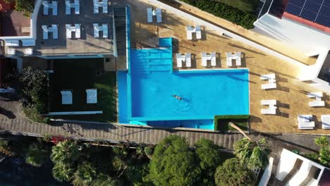 A-young-and-slim-woman-is-swimming-on-her-back,-enjoying-the-cool-water-of-the-pool-during-sunset-in-a-quiet-and-empty-resort-in-Ponta-do-Sol,-Madeira-Island