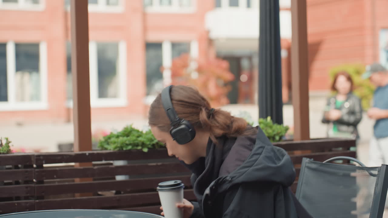 Kvinde på en terrasse, der drikker kaffe med høretelefoner på, sort jakke, takeaway-kop i hånden, afslappet stilling med ansigtet mod gaden, bløde downtown-lyde, weekend-opladning, folk der går forbi i baggrunden
