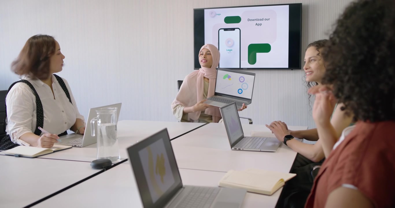 Female coworkers listening as presenter in scarf introducing app on laptop monitor seeking feedback