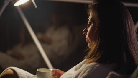 thoughtful woman in bathrobe sits in soft armchair relishing evening coffee. lady with serious facial expression enjoys solitude in tranquil atmosphere