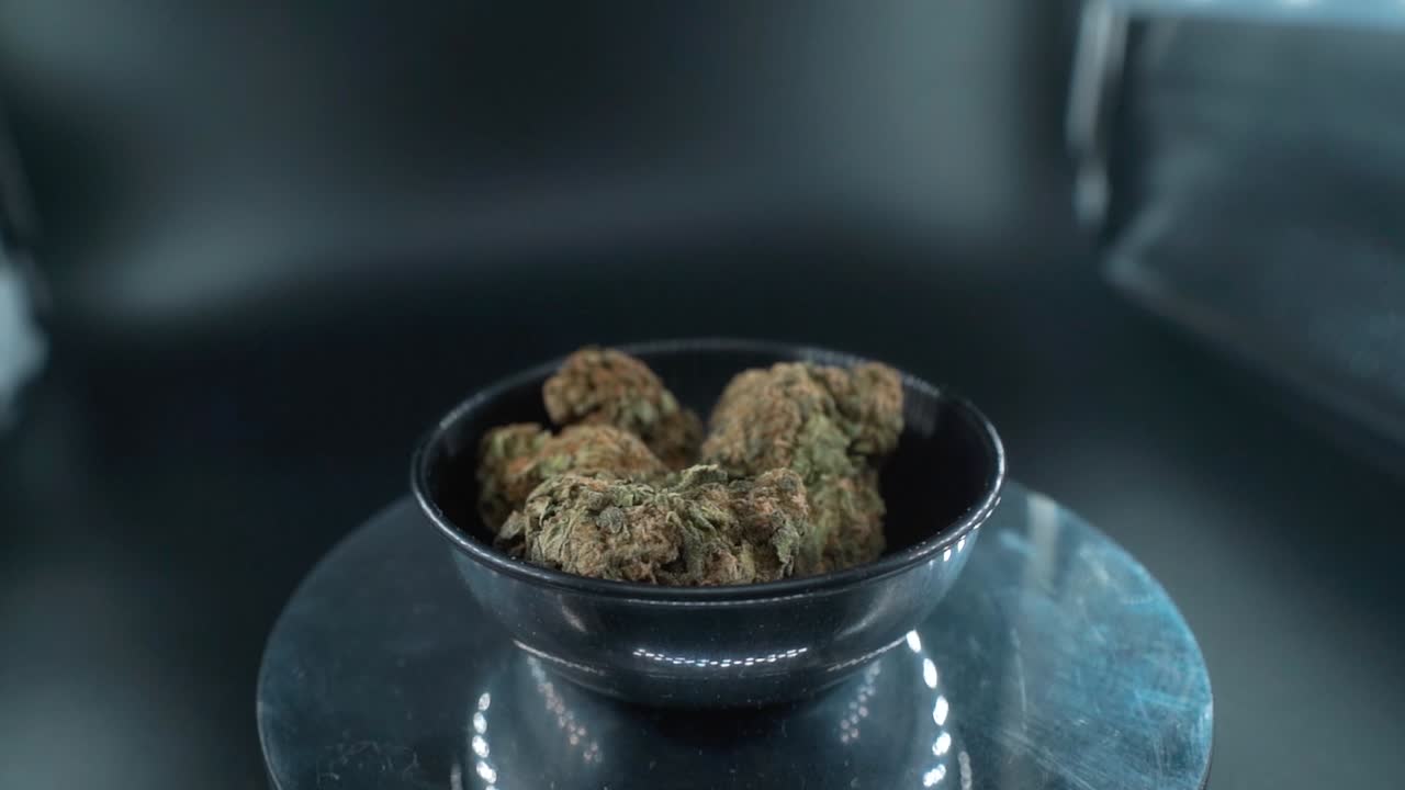 Cime di marijuana essiccate verde-grigiastre - Scatto concettuale ravvicinato, mucchio di piante di marijuana essiccate in una ciotola lucida, ceppi di tricomi, su un supporto rotante riflettente, luci da studio, zoom out lento con gru