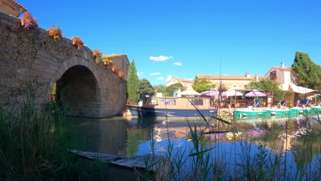 Brücke-über-Den-Kanal-Am-Le-Somail-Canal-Du-Midi-In-Frankreich-Mit-Booten,-Die-An-Einem-Warmen-Sommerabend-Vor-Einer-Bar-Festgemacht-Haben