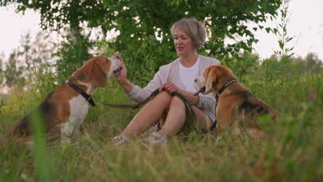 woman seated outdoors on grassy field with her dogs, holding something in her hand while interacting with one dog on her right as she feeds him, while the other dog attentively watches