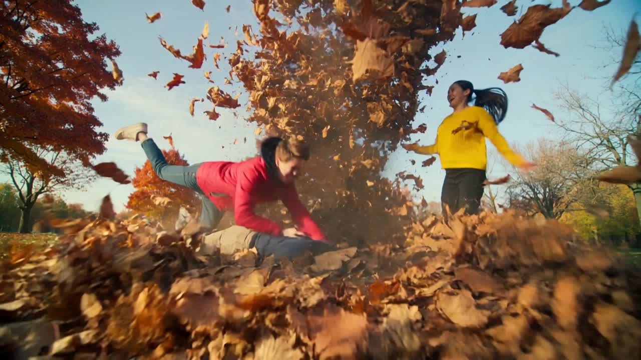 Lunging three adults playing pushing leaf pile in park, red jacket diving, yellow sweater cheering