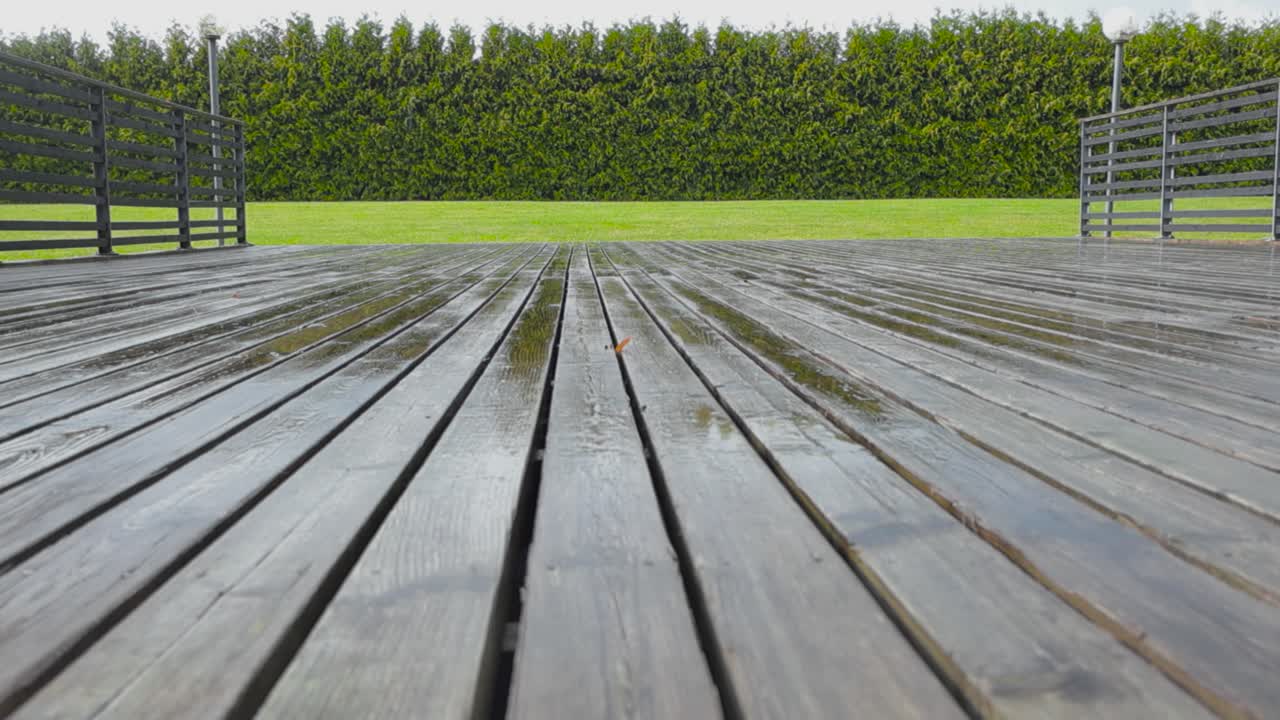 Rain falling on a brown and worn out wooden terrace deck in slow motion during a cloudy autumn or summer day. Water puddles on the terrace are reflective and green short mowed lawn grass in the back.