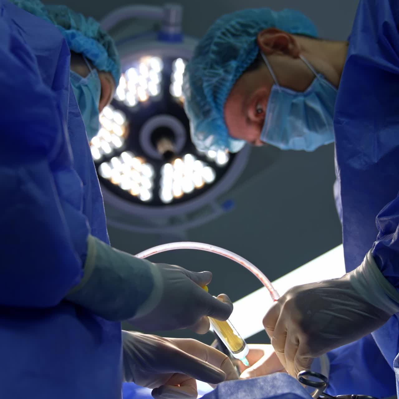 Neurosurgeon uses device with tube while assisting nurse holding a syringe. Medics working under the switched on modern round lamp in surgery room. Low angle view