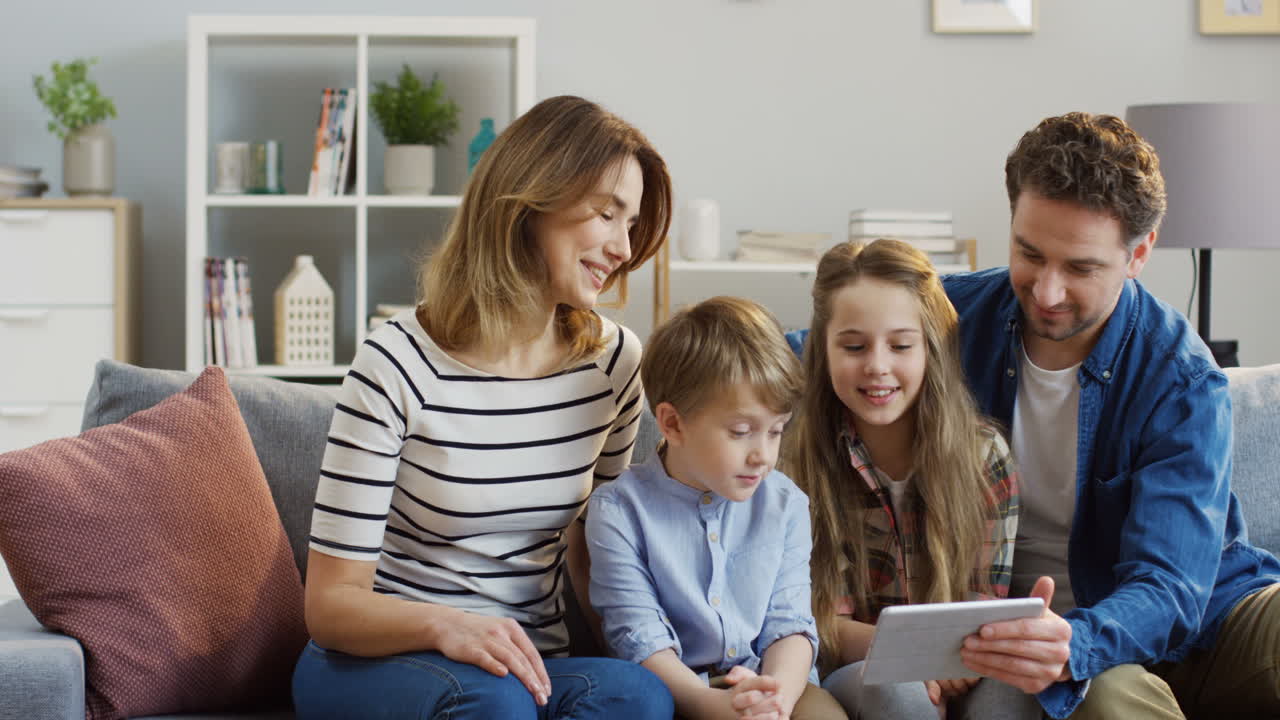 Cheerful Family With Two Kids Sitting On The Sofa, Laughing While Watching Something Very Funny On The Tablet Device