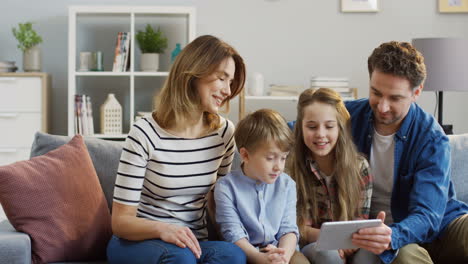 familia alegre con dos niños sentados en el sofá, riéndose mientras ven algo muy divertido en el dispositivo de tableta