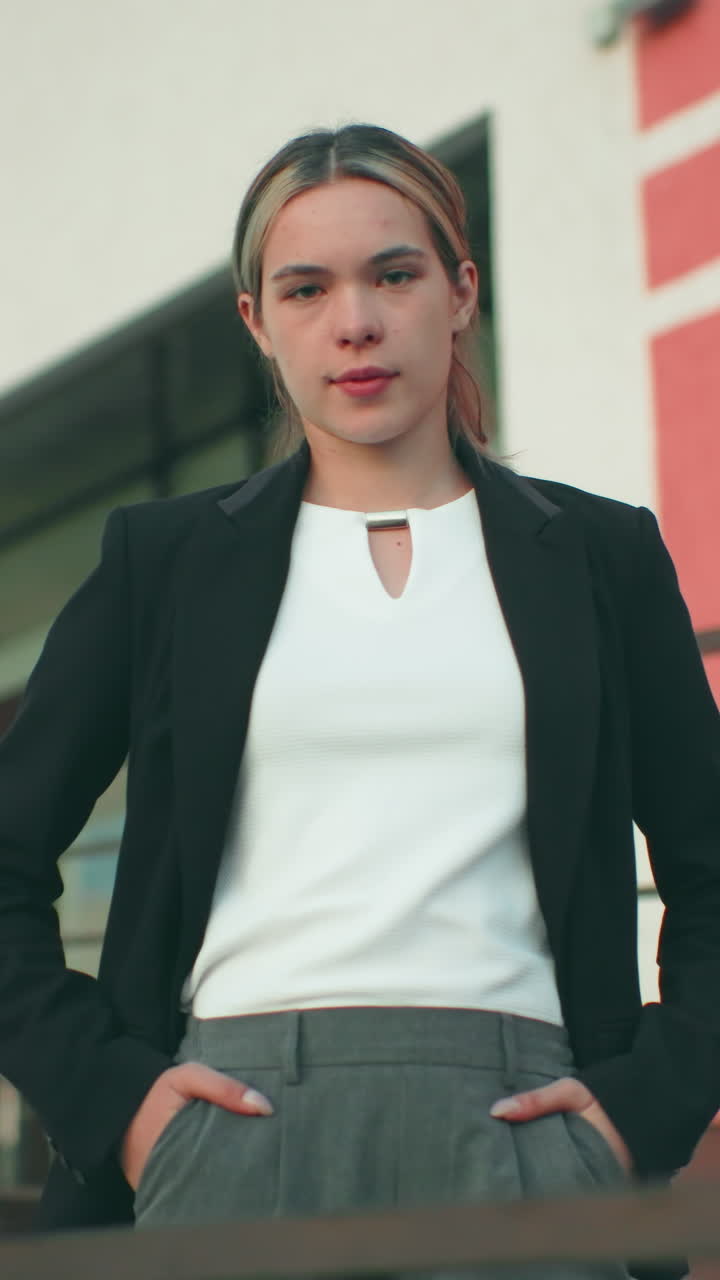 Confident woman in black blazer and white top poses with hands in pockets near iron railing in front of modern office building under clear sky, radiating authority, and professionalism outdoors