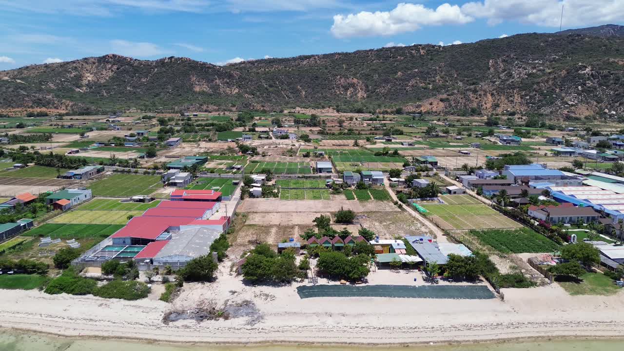 Drone zooming out showcasing the organized farmlands and coastal village near My Hoa Lagoon, with hills in the background under sunny skies.