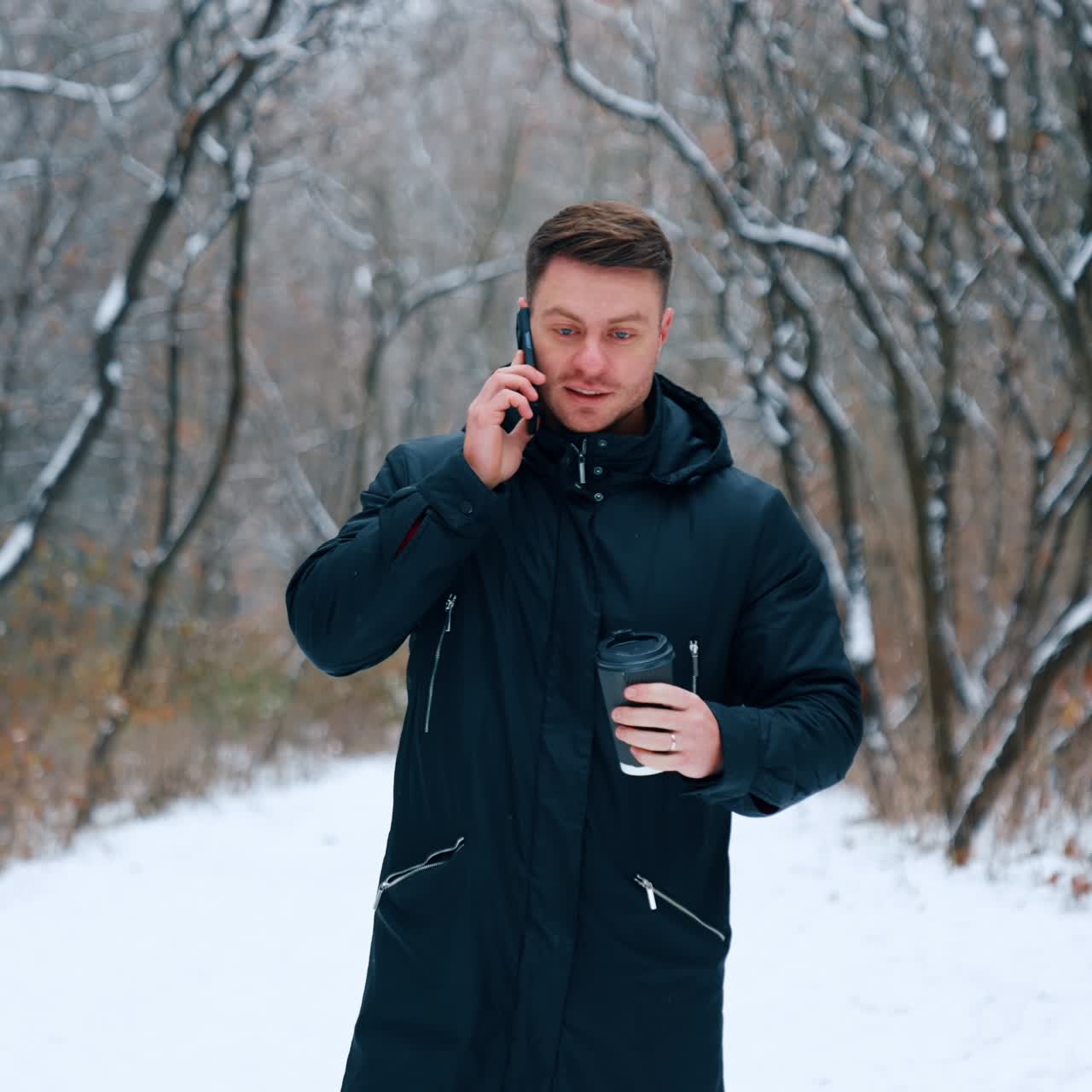Man with coffee cup in hand speaks on the phone. Caucasian male walking by the beautiful park in winter