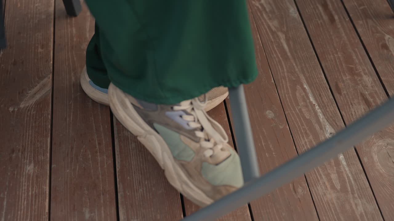 pantaloni verdi, scarpe da ginnastica sotto un tavolo di metallo, momento casual all'aperto che mostra un piede irrequieto che batte e un leggero movimento su una piattaforma di legno, pelle scamosciata usurata e suola graffiata, gamba di sedia in metallo che attraversa l'inquadratura