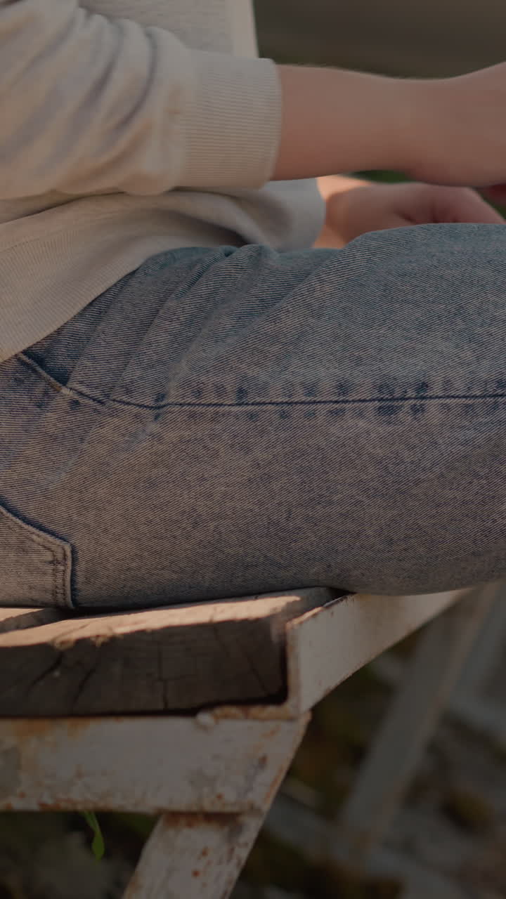 Close-up of woman in jeans sitting on first row of rustic stadium seating, adjusting left leg forward, with hand resting on leg, sunlight and small green plants