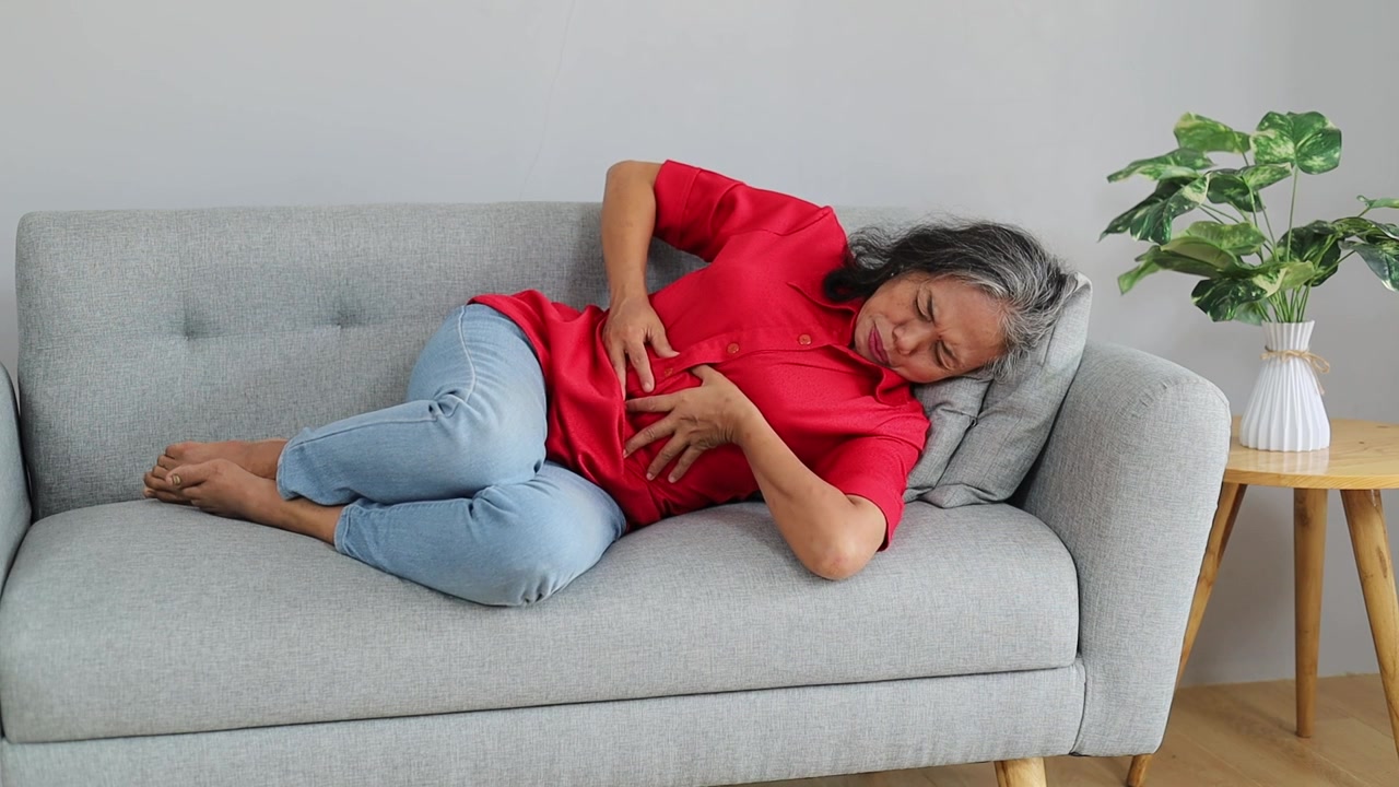Senior Asian woman lying on a sofa and holding her stomach in pain, illustrating abdominal pain, stomachache, and digestive discomfort.