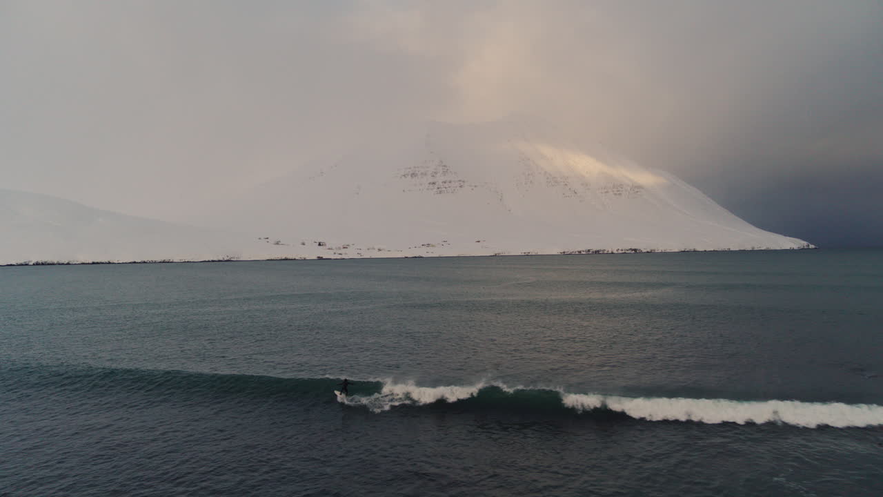 Aerial descend to surfer riding stormy waves battering an Icelandic bay surrounded by dramatic cliffs and icy waters with snow